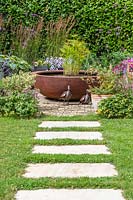 Stepping stone in lawn leading to paved area with corten steel water feature and circular planting