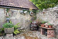 Cobbled farmyard with display of succulents, vintage barrel colonised by Erigeron karvinskianus, galvanised gardenalia and a planted drinking trough 