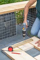 Woman using an electric screwdriver to screw through a batten, to construct a table top made up of cut scaffolding boards. 
