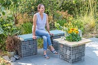Woman sitting on gabion and scaffolding board bench that is complimented by a matching table on slate patio in modern garden. 