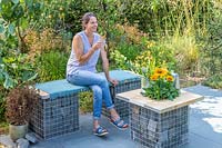 Woman sitting and enjoying a drink on gabion and scaffolding board bench that is complimented by a matching table on slate patio in modern garden. 