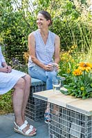 Two women sitting on a gabion bench next to a matching table with built in planter. 