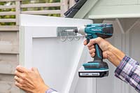 Using a cordless electric drill to attach a set of metal hooks to the inside of a shed door