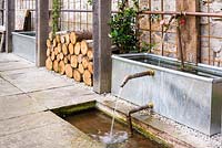 Galvanised drinking trough, copper pipes and taps used as a water feature below a timber pergola in the courtyard garden at Am Brook Meadow, Devon in August.