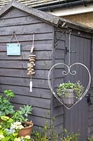 Small wooden grey painted garden shed decorated with a sign, a hanging ornament and a hanging metal basket shaped as a love heart with planted containers below