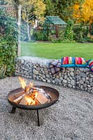 Lit firepit area with colourful cushions on gabion benches and pebble surface, view of garden beyond 