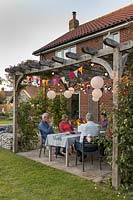People at dining table enjoying snakcs and drinks underneath a wooden pergola decorated with light chains and colourful bunting