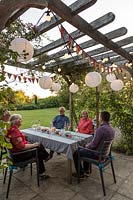 People at dining table enjoying snakcs and drinks underneath a wooden pergola decorated with light chains and colourful bunting
