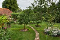 Tiny orchard at the back of the plot catches the early morning sun. With narrow curving brick pathway. and wooden plant signs. Trees include an almond, prunus dulcis, surrounded by white dianthus, as well as apples and pears.