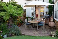 Small garden patio with table and parasol, barbecues and Dicksonia antarctica - Tree Fern - in bed near lights