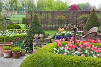 Relaxing area on paved courtyard with displays of spring flowers in containers, bordered with box hedging and yew topiary.