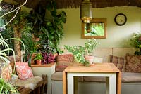 Jungle Hut constructed using recycled telegraph poles with cushioned benches, mirror, clock, lamp and table surrounded by houseplants including begonias, tradescantias, clivias and spider plants.