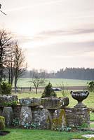 Low stone wall displayed with troughs and classic urns, with view to countryside beyond at Rodmarton Manor, Glos, UK. 