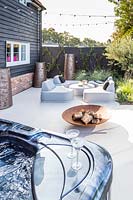 Paved area with modern lounge furniture and firepit, jacuzzi in foreground. 