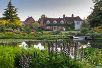 View over the pond to the pontoon and house. Planting includes Acanthus mollis, waterlilies, yellow loosestrife and Lysimachia punctata, in front of the herbaceous border on the other side.