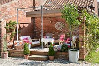 Raised seating area with a metal pergola and a shade cover against side of a brick wall, decorated with lounge furniture and pots