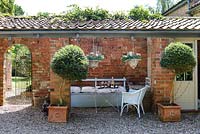 Covered seating area with wooden painted bench and table under overhang, standard trees in pots and hanging baskets 
