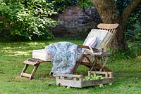 Wooden deckchair lounger with cushions and blanket on a lawn in the shade under a tree 