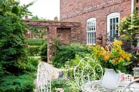 Small courtyard with decorative white metal furniture and vase with flowers, foliage bed against brick house and wall