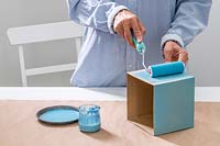 Woman using a small roller to paint a cardnoard box with blue paint