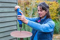 Woman using electic screwdriver to fix wooden disk to post. 