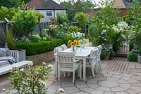 Table and chairs in paved dining area in small town garden, with white-themed borders. 