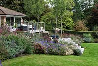 Autumn borders of herbaceous flowering plants and ornamental grasses wrap around a contemporary deck and terrace, linked by steps. Four 'Chanticleer' ornamental pear trees are planted in lowest terrace.