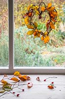 Simple heart shaped willow wreath with autumnal Hornbeam foliage and fairy lights hanging in shed window with view to garden