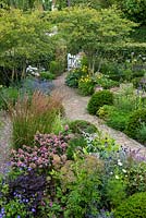 A contemporary garden design, a brick path cuts diagonally through the garden, flanked by multi-stemmed Amelanchier lamarckii, in front a bed with ornamental grasses, Escallonia, Taxus - Yew - topiary