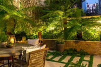 A walled town courtyard with outdoor dining lit at night, illuminating Dicksonia antarctica - Tree fern, Hydrangea and Hosta in raised beds