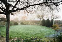 Circular lawn surrounded by borders, clipped yews and Crataegus x lavalleei 'Carrierei' on a misty March morning
