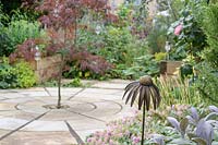 Metal flower head in a border, beyond circular paving with centrally planted Acer - Maple
