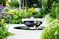 View of David Harber water feature surrounded by Allium 'Globemaster', Luzula nivea and Euphorbia characias subsp. wulfenii. 