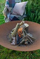 A lit fire bowl made from rusted corten steel, a wooden garden chair with a wool rug and a cushion by a large rosemary bush.