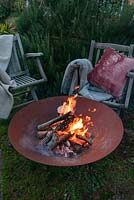 A lit fire bowl made from rusted corten steel,  wooden garden chairs with wool rugs and a cushion by a large rosemary bush.