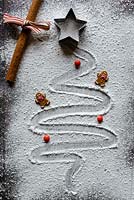 A Christmas tree drawn in icing sugar on a wooden chopping board with a star cutter, ginger bread men, red chocolate buttons and a cinnamon stick tied with ribbon.