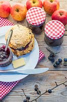 Scone filled with Sloe Berry Jam on table top with jars of jam, apples, sloe berries and gingham table cloth. 