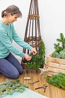 Woman using brown garden twine to attach pieces of pine - abies foliage to willow obelisk frame