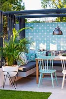Dining area with colourful wall, chairs, modern pergola, industrial style lamp and the palm in the white pot.