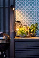 Illuminated worktop with ferns planted in small pots and lanterns by colorful wall in an outdoor kitchen at night.