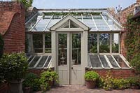 Small wooden greenhouse set in brick walls in courtyard garden, with cold frames with tender plants growing inside. 