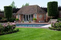 Swimming pool and pool house in walled garden with brick paving, edged with large topiary columns and  herbaceous borders filled with pink roses.