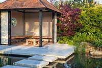 Garden pavilion with wooden benches approached by floating stone stepping stones over a pond, plants: Acer palmatum 'Bloodgood' - Japanese Maple, ferns and Equisetum hyemale. A Japanese Reflection Garden 