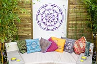 An outdoor sofa with candles and colourful cushions from India, with a lotus and ohm panel screen, wooden fence and bamboo plants. Relaxation, Meditation garden, RHS Tatton Park Flower Show, 2017. Designer: Paul Morris and Mike Ball. 