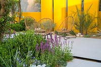 Mediterranean climate garden grey stone paved patio seating area with table and cane chairs - yellow screen wall a rill and planting of Salvia nemorosa in the foreground, dry gravel bed with cacti, aloes and yucca plants in the background  Santa Rita Living La Vida 120 Garden. RHS Hampton Court Flower Show July 2018 - Designer: Alan Rudden - Sponsor Santa Rita wine 