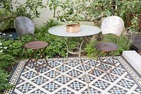 Metal table and chairs on a tiled mosaic patio, behind bed with Malus - Apple - trees and underplanting.The Style and Design Garden, sponsors CED Stone, London Mosaic, Garden Brocante Online