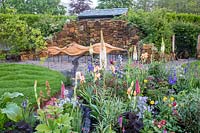 Small flower bed planted with Irises, Eremurus, delphiniums, lupins, Primula viallii - carved oak wood bench on gravel seating area - small grass lawn. Grace and Dignity garden. RHS Malvern Spring Festival May 2019 -  Designer Lucie Giselle Ponsford - 