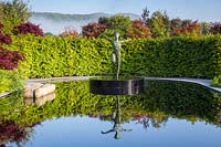 Reflective black pond water feature with sculpture 'Zephyr' by Simon Gudgeon on a misty May spring day surrounded by garden hedge and acers. Garden of Quiet Contemplation garden.  RHS Malvern Spring Festival May 2019 - Designer Peter Dowle - Leaf Creative 