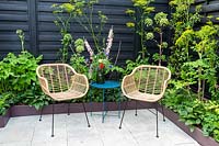 Stone paved patio and garden furniture metal blue table and with rattan metal framed chairs - painted black garden fence and small raised bed borders with Angelica - Hostas and ferns in a mainly green colour scheme