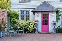 A contemporary country cottage front garden planted with Hydrangea 'Limelight, patio seating area and urns with annual geraniums.